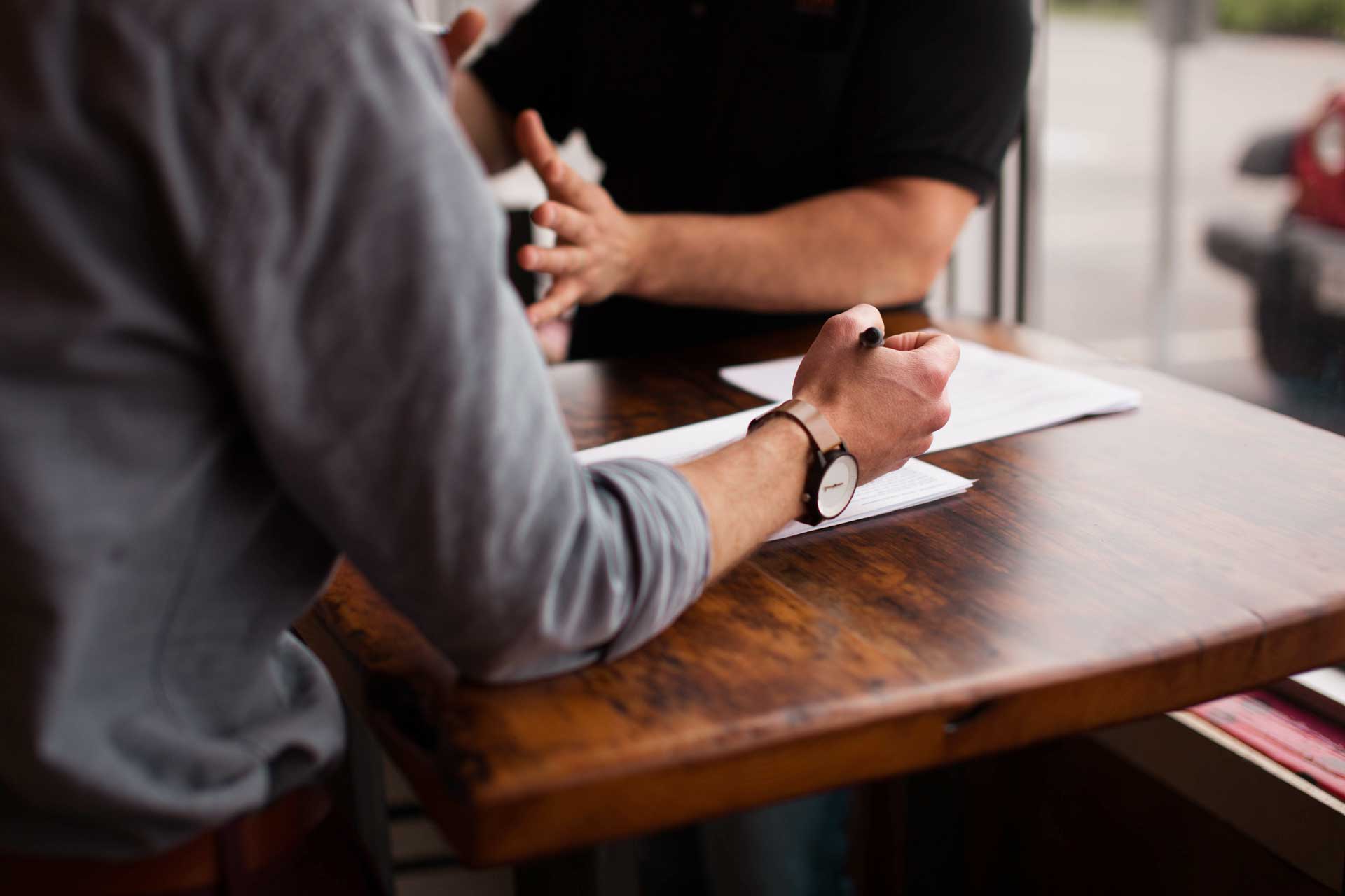 men sitting at wooden desk working with pen paper and gesturing with their hands