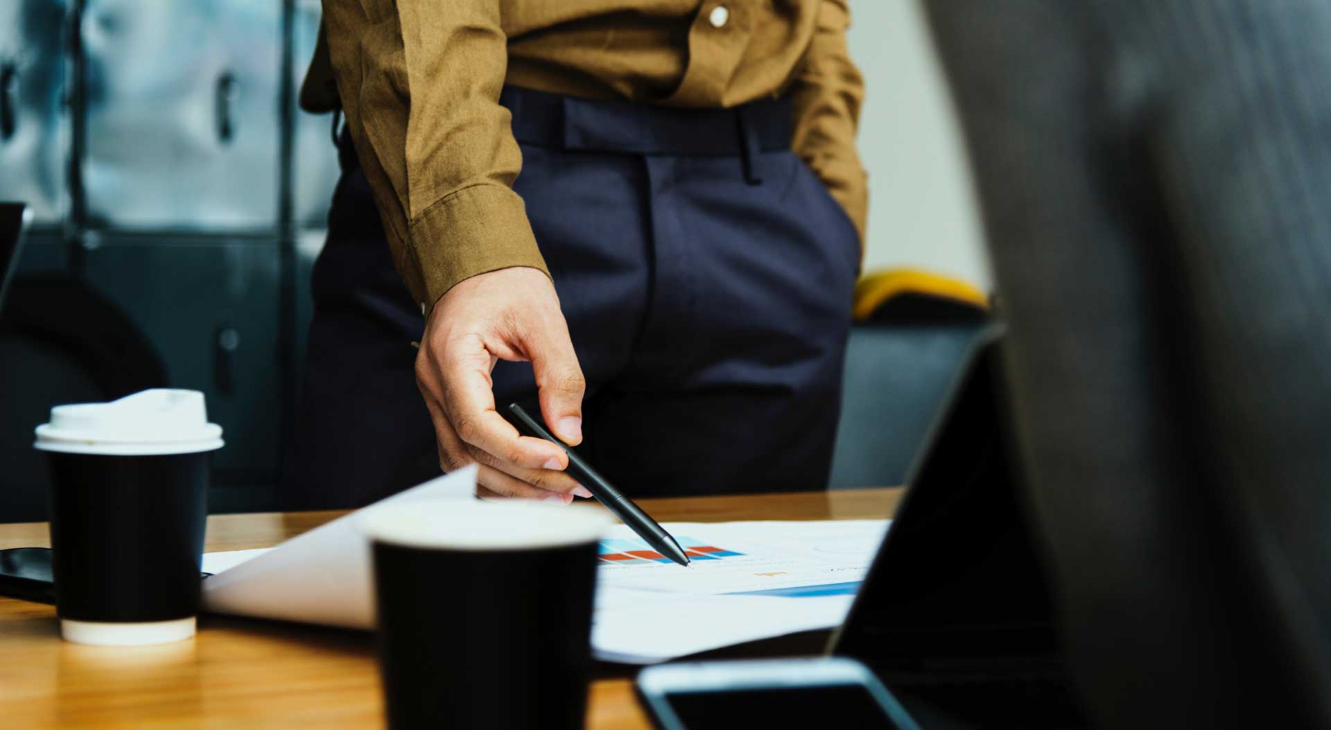 standing man in formal business attire pointing with pen at documents on able